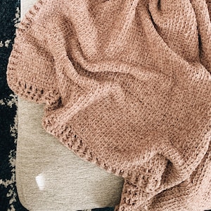 Może przedstawiać: Miękki, różowy pled szydełkowy drapowany na jasnoszarym pufie.