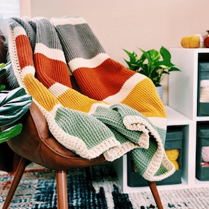 Könnte beinhalten: Eine mehrfarbige gestreifte gehäkelte Wolldecke, die über einem braunen Ledersessel mit Holzbeinen drapiert ist. Die Decke hat Streifen in Grau, Weiß, Orange, Gelb und Grün.