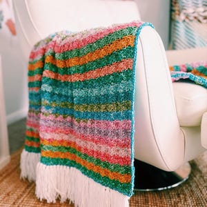May include: A vibrant, striped crochet blanket with a white fringe is draped over a white chair. The blanket displays horizontal stripes in shades of pink, orange, green, and blue. The chair has a modern design.