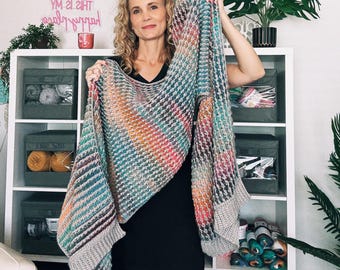 May include: A colorful, handmade shawl with a houndstooth pattern. The shawl features a gradient of colors including teal, orange, pink, and gray. The edges are finished with a solid gray border. The shawl is held up in front of a white shelf.