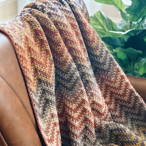 Pu&ograve; includere: Una coperta all'uncinetto con un motivo a chevron in tonalit&agrave; di marrone, arancione e giallo. La coperta &egrave; drappeggiata su una poltrona in pelle marrone.