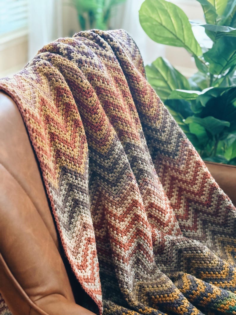 Pu&ograve; includere: Una coperta all'uncinetto con un motivo a chevron in tonalit&agrave; di marrone, arancione e giallo. La coperta &egrave; drappeggiata su una poltrona in pelle marrone.
