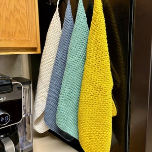 K&ouml;nnte beinhalten: Vier geh&auml;kelte Geschirrt&uuml;cher h&auml;ngen an Haken. Die T&uuml;cher sind in den Farben beige, blau, gr&uuml;n und gelb.