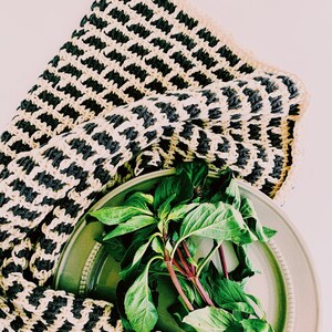 May include: A green ceramic plate with a bunch of fresh green basil leaves on top. A black and white striped crocheted dishcloth is draped over the plate.