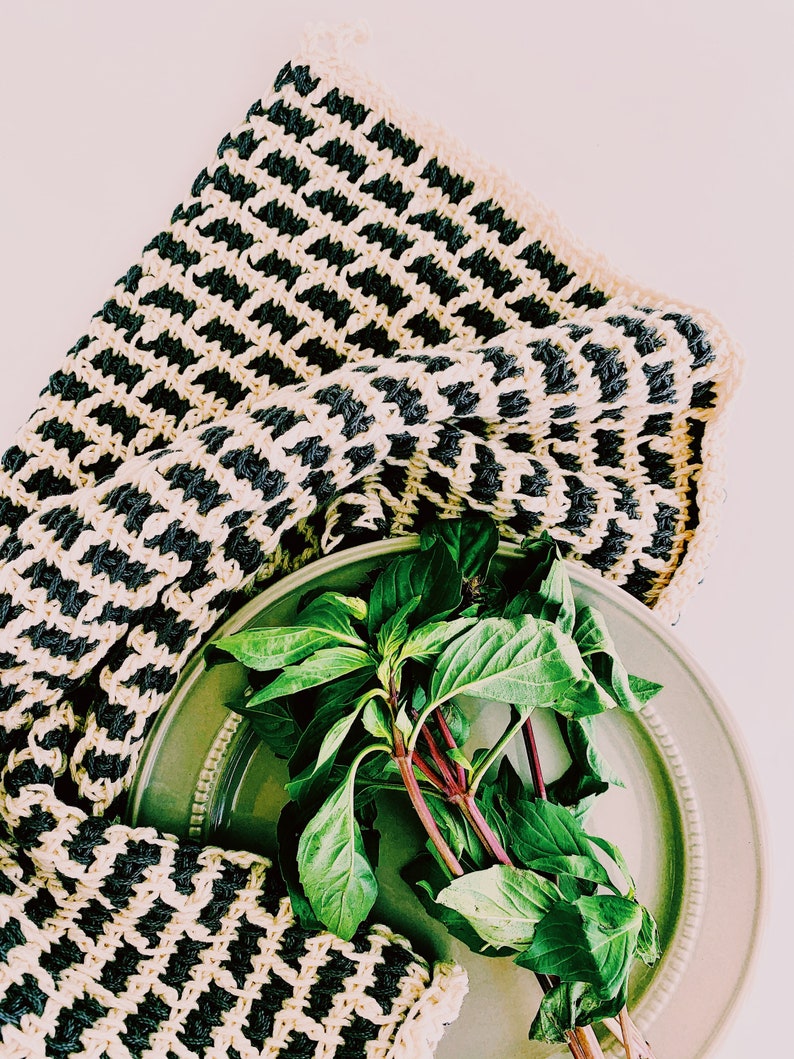 May include: A green ceramic plate with a bunch of fresh green basil leaves on top. A black and white striped crocheted dishcloth is draped over the plate.