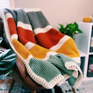 May include: A multi-colored striped crocheted throw blanket in shades of gray, white, orange, yellow, and green. The blanket is draped over a brown leather armchair.