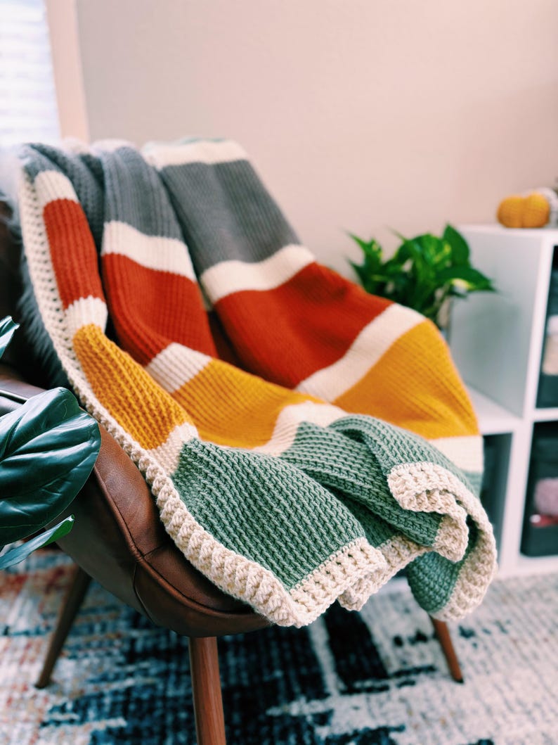 May include: A multi-colored striped crocheted throw blanket in shades of gray, white, orange, yellow, and green. The blanket is draped over a brown leather armchair.