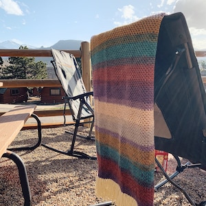 Pu&ograve; includere: Una coperta a righe in crochet con frange appesa a una sedia pieghevole nera. La sedia &egrave; su un patio con un tavolo di legno e una vista sulle montagne in lontananza.