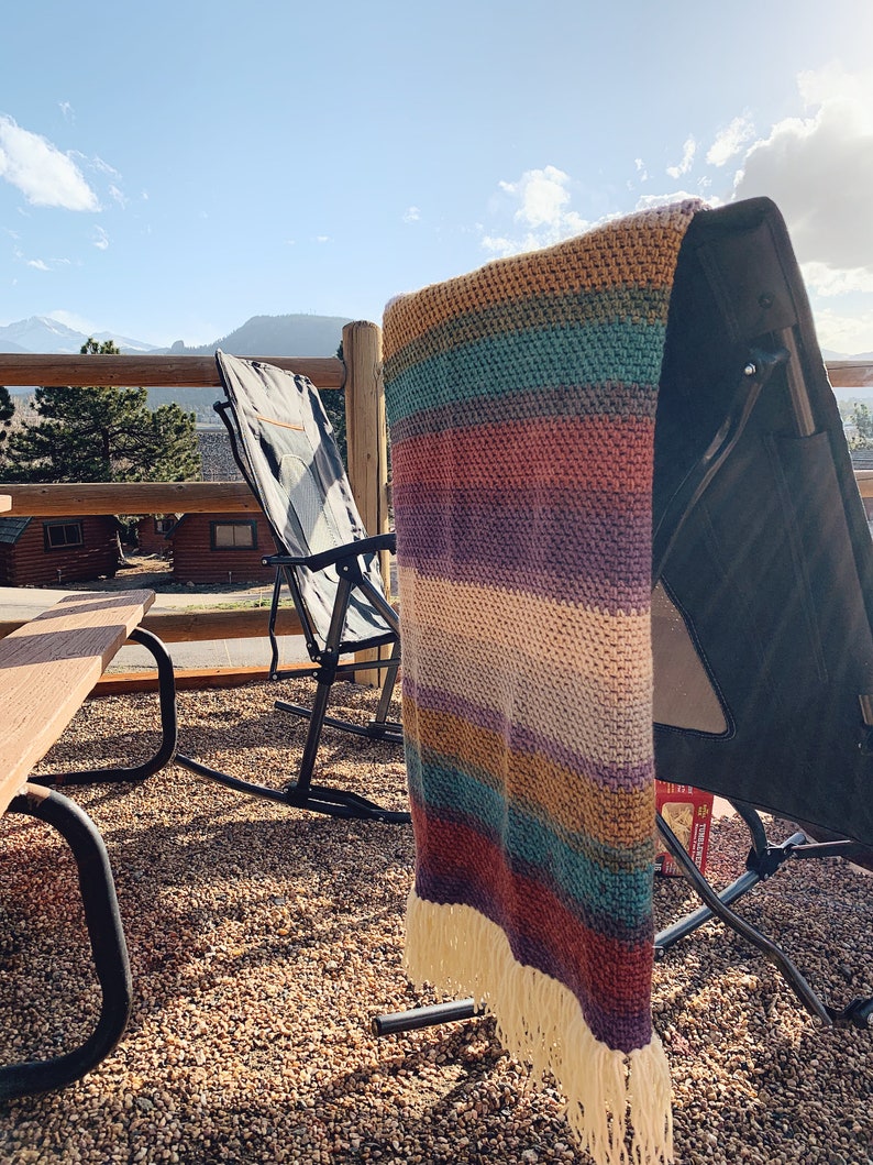 Pu&ograve; includere: Una coperta a righe in crochet con frange appesa a una sedia pieghevole nera. La sedia &egrave; su un patio con un tavolo di legno e una vista sulle montagne in lontananza.