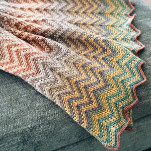 Pu&ograve; includere: Una coperta all'uncinetto multicolore con un motivo a chevron. La coperta &egrave; realizzata con una variet&agrave; di tonalit&agrave; di marrone, arancio, giallo, verde e blu. La coperta &egrave; piegata e drappeggiata su un divano grigio.
