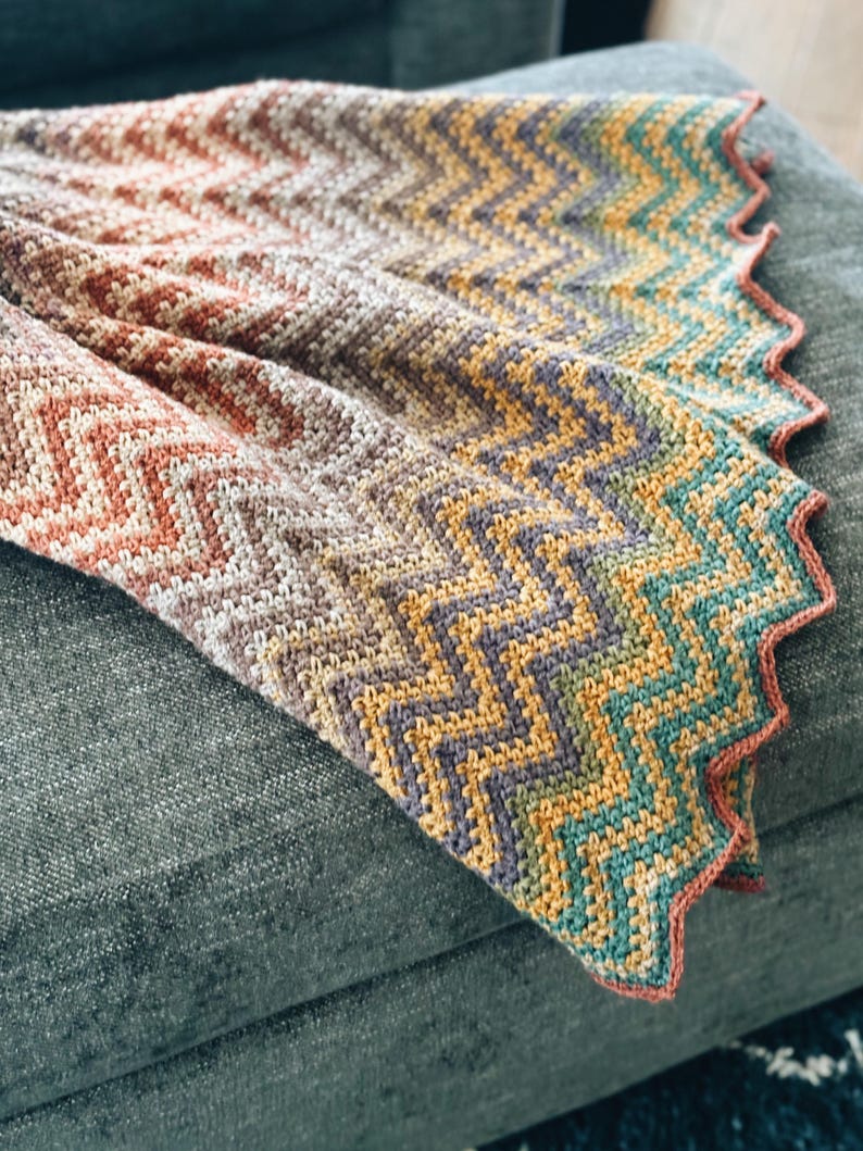 Pu&ograve; includere: Una coperta all'uncinetto multicolore con un motivo a chevron. La coperta &egrave; realizzata con una variet&agrave; di tonalit&agrave; di marrone, arancio, giallo, verde e blu. La coperta &egrave; piegata e drappeggiata su un divano grigio.
