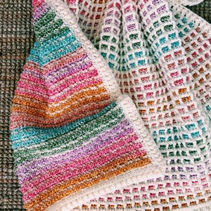 Op de afbeelding: Een handgemaakte gehaakte deken met een levendig regenboogverloop. De deken heeft een mix van kleuren, waaronder roze, oranje, groen en blauw, met een witte rand. Het open weefpatroon voegt textuur en visuele interesse toe.