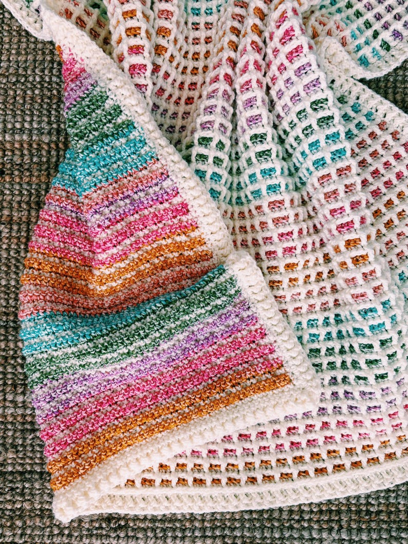 Op de afbeelding: Een handgemaakte gehaakte deken met een levendig regenboogverloop. De deken heeft een mix van kleuren, waaronder roze, oranje, groen en blauw, met een witte rand. Het open weefpatroon voegt textuur en visuele interesse toe.