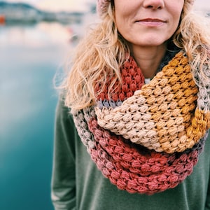 May include: A woman wearing a multi-colored crocheted infinity scarf with a pink beanie.