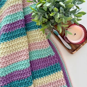 Pode incluir: Um cobertor de crochê feito à mão em cores pastel, com listras em turquesa, lavanda, rosa e creme. O cobertor tem um padrão de ponto texturizado e é dobrado. Uma pequena planta, vela e agulha de crochê estão no fundo.