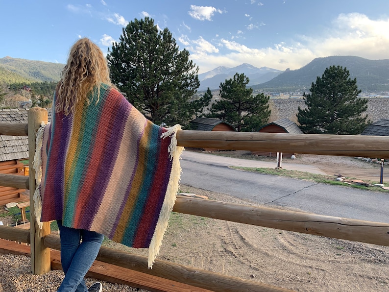 Pu&ograve; includere: Una persona indossa un poncho a righe colorate in crochet con frange, in piedi su un ponte di legno con vista su una catena montuosa.