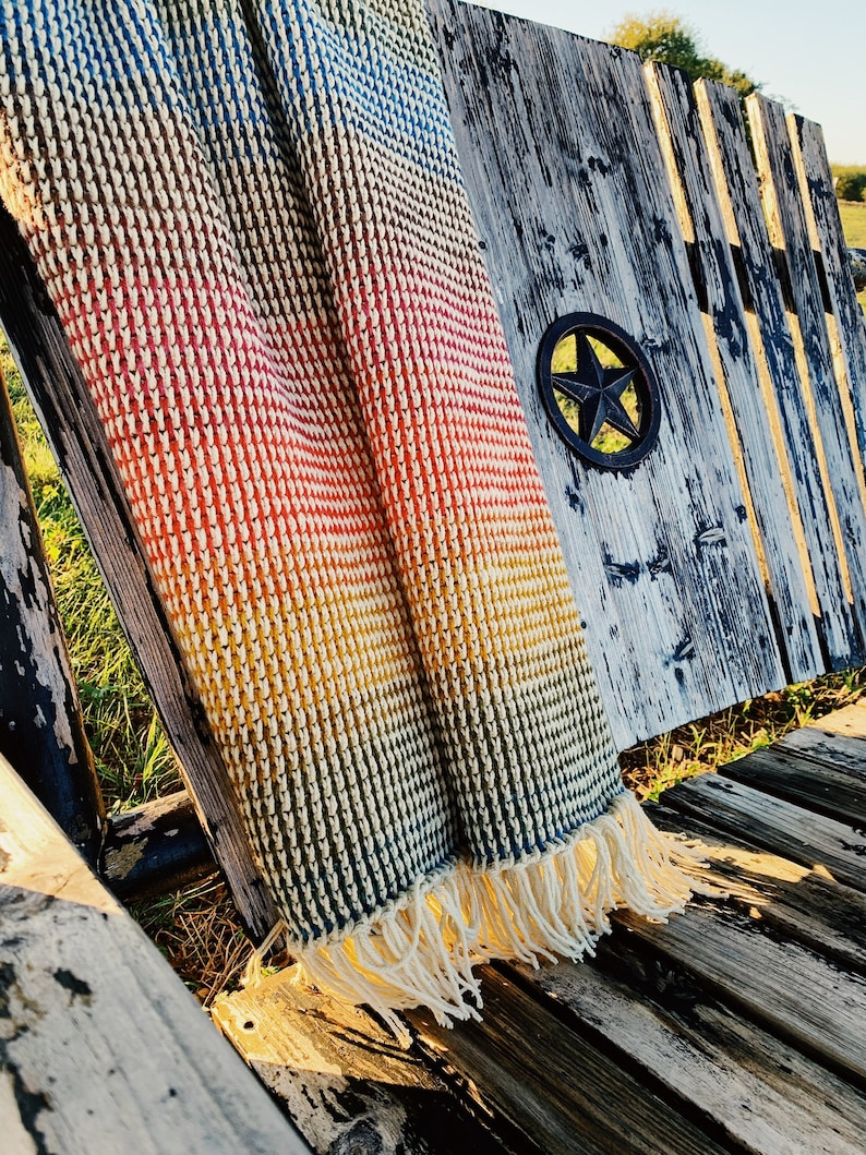 Pu&ograve; includere: Una coperta di uncinetto color arcobaleno con frange che pendono dal bordo. La coperta &egrave; drappeggiata su una panca di legno con una stella di metallo sul retro.