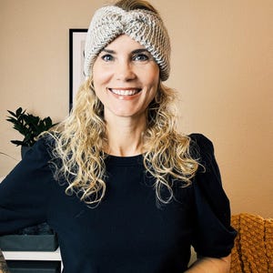 Può includere: Una donna con i capelli biondi ricci indossa una fascia per capelli in maglia grigia e bianca con un nodo attorcigliato in cima. Sta sorridendo e guardando la camera. Indossa una camicia nera con maniche a sbuffo.