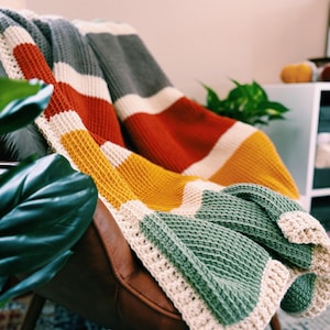May include: A multi-colored striped crocheted throw blanket in shades of gray, white, orange, yellow, and green. The blanket is draped over a brown leather armchair.