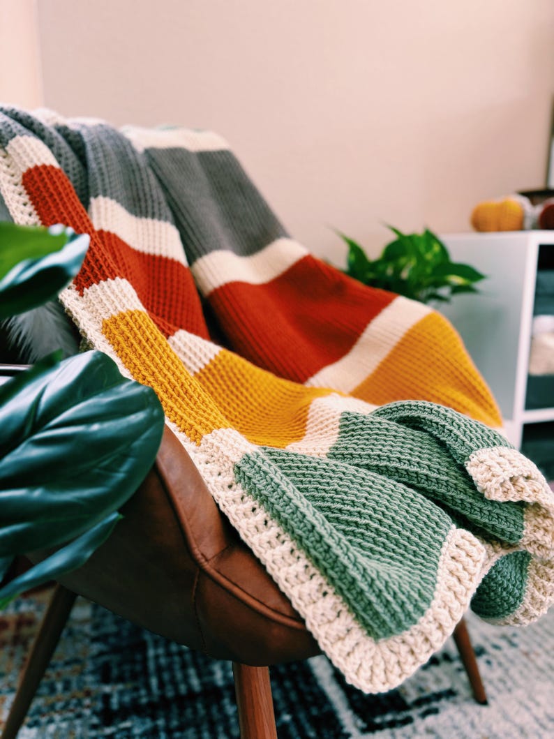 May include: A multi-colored striped crocheted throw blanket in shades of gray, white, orange, yellow, and green. The blanket is draped over a brown leather armchair.