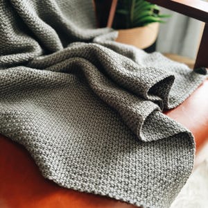 Op de afbeelding: Een grijze gebreide deken gedrapeerd over een bruine leren fauteuil. De deken heeft een textuur, een wafelijzerpatroon.