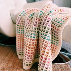 Op de afbeelding: Een cr&egrave;mekleurige, gehaakte deken met een rasterpatroon. De deken heeft een regenboog aan kleuren, waaronder roze, oranje, groen en blauw. De deken is over een witte stoel gedrapeerd, wat een gezellige en uitnodigende sc&egrave;ne cre&euml;ert.