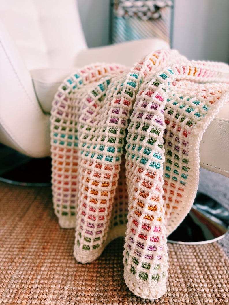 Op de afbeelding: Een cr&egrave;mekleurige, gehaakte deken met een rasterpatroon. De deken heeft een regenboog aan kleuren, waaronder roze, oranje, groen en blauw. De deken is over een witte stoel gedrapeerd, wat een gezellige en uitnodigende sc&egrave;ne cre&euml;ert.