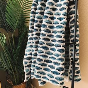 Puede incluir: Una manta de crochet azul y blanca con un patrón geométrico. La manta está colgada sobre una escalera de metal negro.