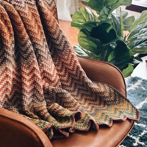 Pu&ograve; includere: Una coperta all'uncinetto con un motivo a chevron in tonalit&agrave; di marrone, arancione e verde. La coperta &egrave; drappeggiata su una poltrona in pelle marrone.