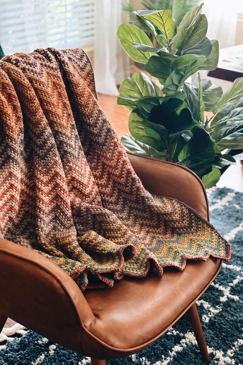Pu&ograve; includere: Una coperta all'uncinetto con un motivo a chevron in tonalit&agrave; di marrone, arancione e verde. La coperta &egrave; drappeggiata su una poltrona in pelle marrone.