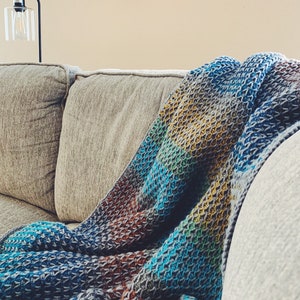 Puede incluir: Una manta de punto de ganchillo multicolor drapeada sobre un sof&aacute; gris. La manta presenta un patr&oacute;n de punto texturizado y un degradado de azul, verde, amarillo y marr&oacute;n.