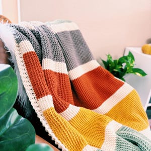 May include: A striped crocheted throw blanket in shades of gray, white, orange, yellow, and green. The blanket is folded and draped over a chair.