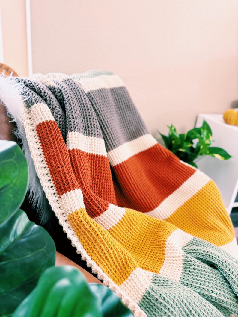 May include: A striped crocheted throw blanket in shades of gray, white, orange, yellow, and green. The blanket is folded and draped over a chair.