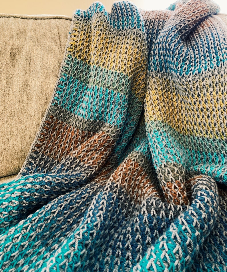 Puede incluir: Una manta de crochet multicolor con rayas de color turquesa, marr&oacute;n y amarillo. La manta est&aacute; doblada y drapeada sobre un sof&aacute;.