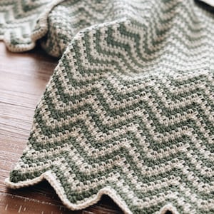 Op de afbeelding: Een gehaakt deken in groen en wit met een gegolfde rand. Het deken is gemaakt met een chevron-patroon.