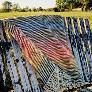 Pu&ograve; includere: Una coperta in crochet a righe colorate con frange, drappeggiata su una panca di legno invecchiato.