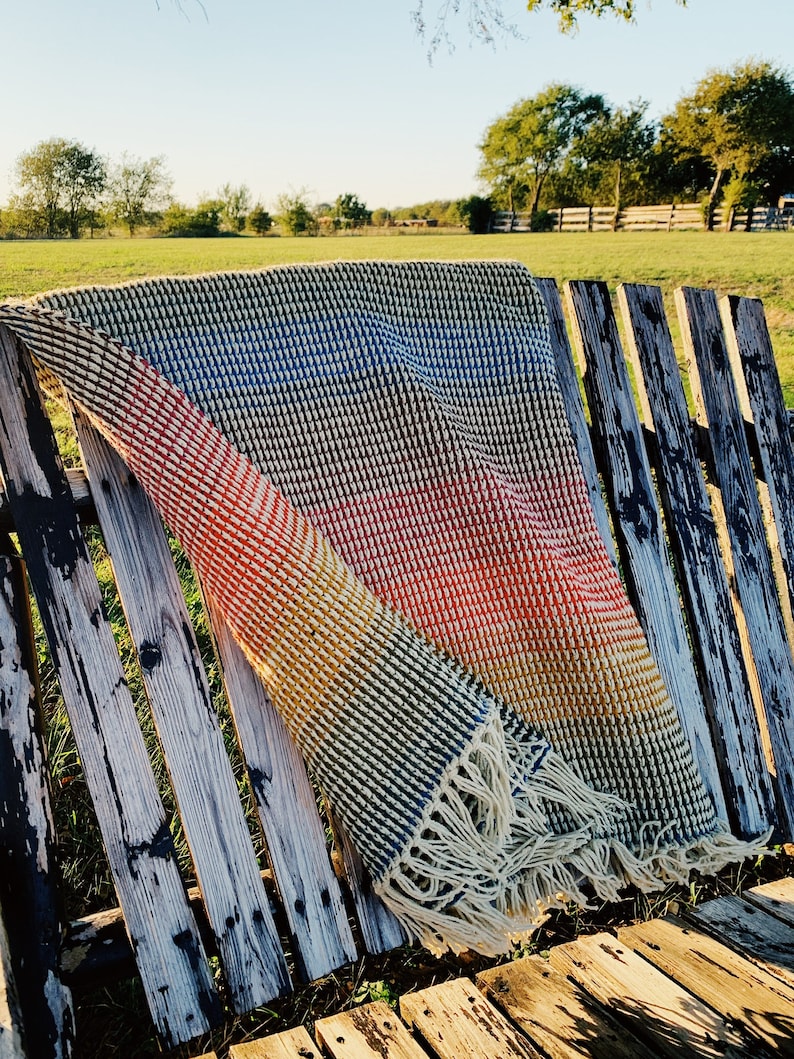 Pu&ograve; includere: Una coperta in crochet a righe colorate con frange, drappeggiata su una panca di legno invecchiato.