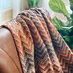 Pu&ograve; includere: Una coperta all'uncinetto con un motivo a chevron in tonalit&agrave; di marrone, arancione e viola. La coperta &egrave; drappeggiata su una poltrona in pelle marrone.