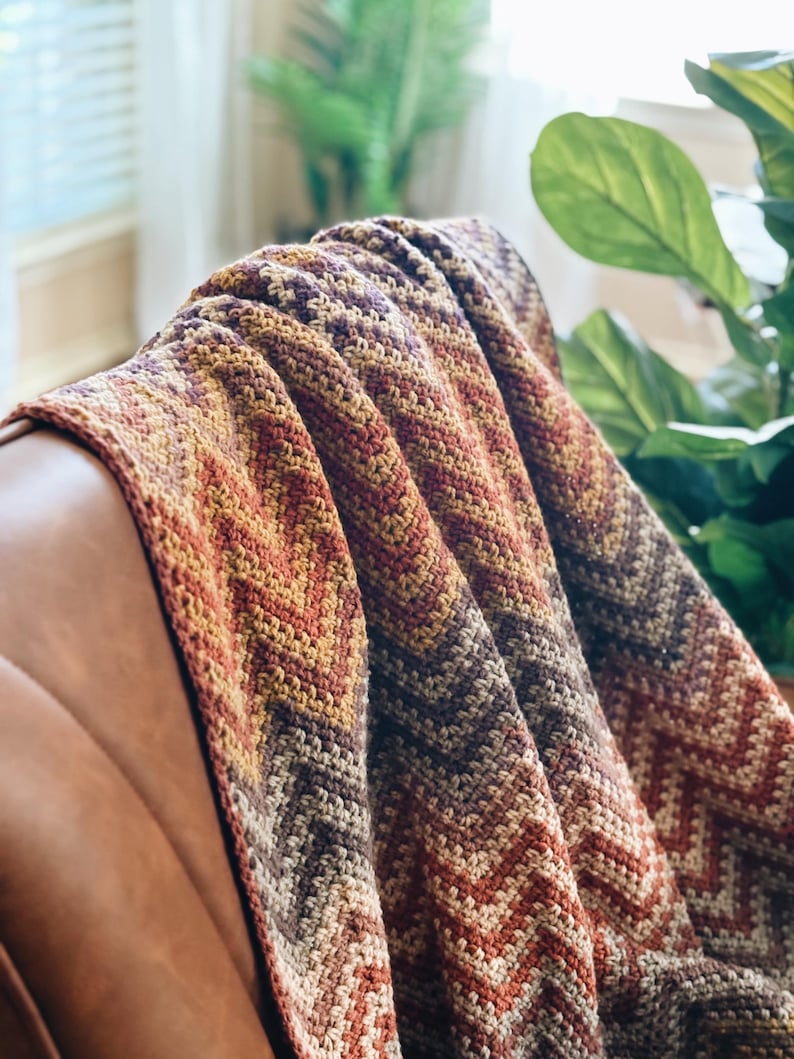 Pu&ograve; includere: Una coperta all'uncinetto con un motivo a chevron in tonalit&agrave; di marrone, arancione e viola. La coperta &egrave; drappeggiata su una poltrona in pelle marrone.