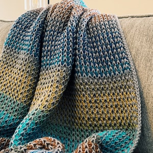 Puede incluir: Una manta de punto de ganchillo multicolor con rayas de color turquesa, marr&oacute;n, amarillo y gris. La manta est&aacute; doblada y drapeada sobre un sof&aacute;.