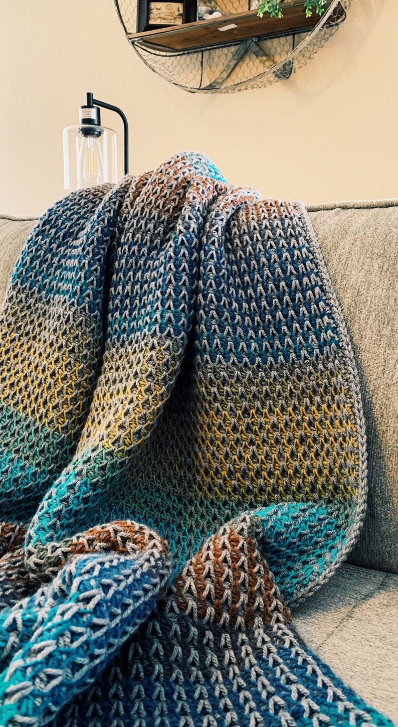 Puede incluir: Una manta de punto de ganchillo multicolor con rayas de color turquesa, marr&oacute;n, amarillo y gris. La manta est&aacute; doblada y drapeada sobre un sof&aacute;.