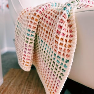 Op de afbeelding: Een handgemaakte, kleurrijke gehaakte deken over een witte stoel. De deken heeft een rasterpatroon met een kleurengradi&euml;nt, waaronder roze, oranje, groen en blauw. De zachte textuur en levendige tinten cre&euml;ren een gezellige en uitnodigende uitstraling.