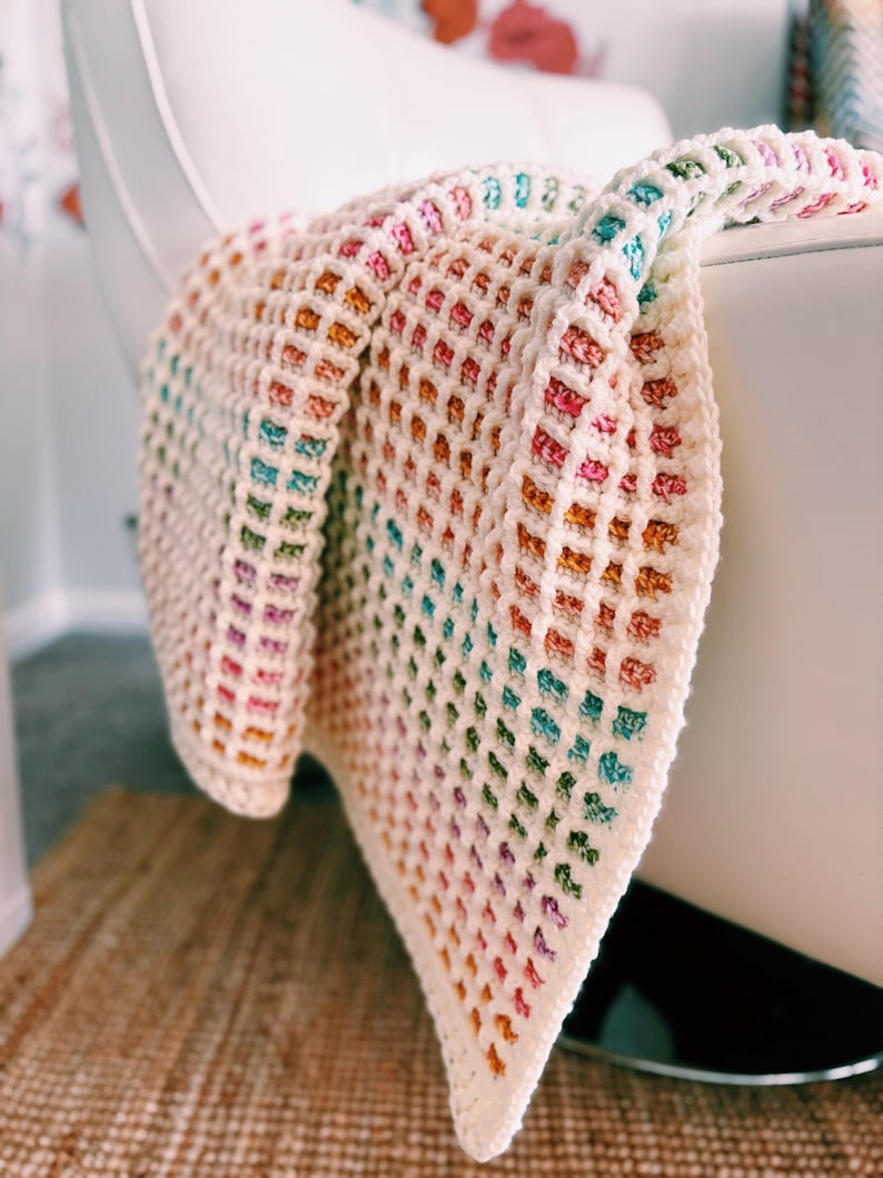 Op de afbeelding: Een handgemaakte, kleurrijke gehaakte deken over een witte stoel. De deken heeft een rasterpatroon met een kleurengradi&euml;nt, waaronder roze, oranje, groen en blauw. De zachte textuur en levendige tinten cre&euml;ren een gezellige en uitnodigende uitstraling.