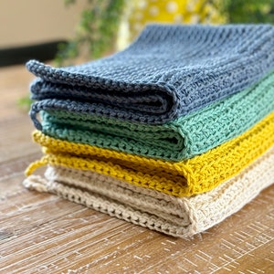 May include: A stack of four crocheted dishcloths in various colors, including blue, green, yellow, and white. The dishcloths are folded and stacked on a wooden surface.