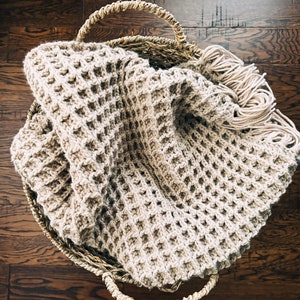 May include: A beige crocheted throw blanket with fringe, folded and placed in a woven basket.