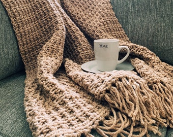 May include: A tan crocheted throw blanket with fringe on a gray couch. A white mug with the word "Mug" printed on it is sitting on a white saucer on top of the blanket.