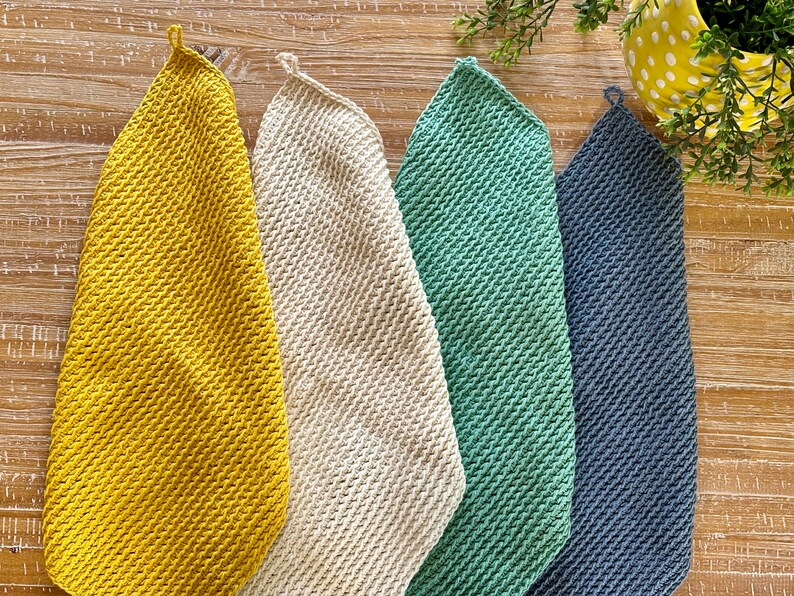 K&ouml;nnte beinhalten: F&uuml;nf geh&auml;kelte Geschirrt&uuml;cher in verschiedenen Farben: Gelb, Wei&szlig;, Gr&uuml;n und Blau. Die Geschirrt&uuml;cher h&auml;ngen an einer Holzoberfl&auml;che.