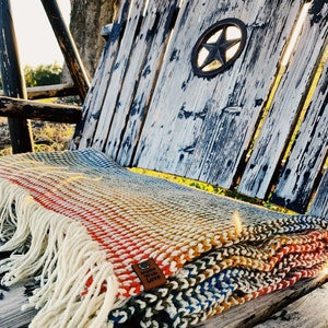 Pu&ograve; includere: Una coperta di maglia multicolore con frange su una panca di legno. La coperta ha un'etichetta in pelle marrone che dice "Made with Love".