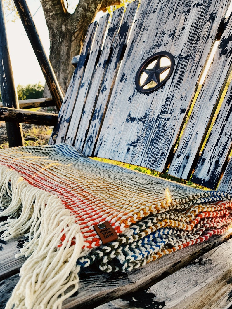Pu&ograve; includere: Una coperta di maglia multicolore con frange su una panca di legno. La coperta ha un'etichetta in pelle marrone che dice "Made with Love".