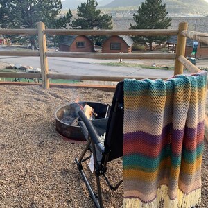 Pu&ograve; includere: Una coperta all'uncinetto a righe con frange &egrave; appesa a una sedia pieghevole nera davanti a un focolare. La sedia &egrave; posizionata su una superficie di ghiaia con vista su una catena montuosa e cabine in lontananza.
